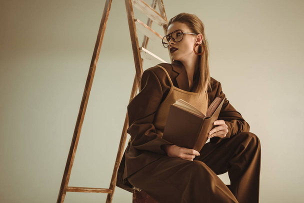beautiful stylish girl holding book and sitting near ladder isolated on beige - Photo, Image