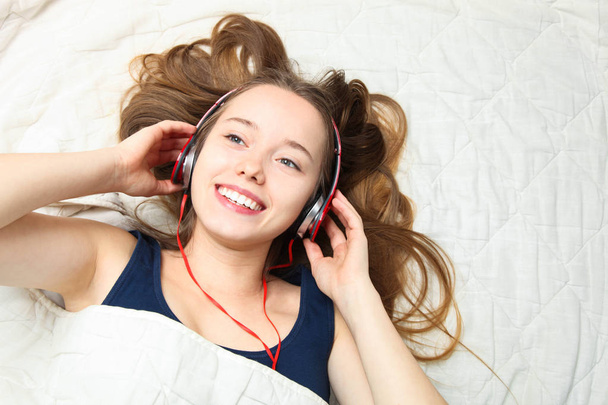 Joven hermosa chica caucásica feliz mienten y escucha música con auriculares en la cama blanca. Concepto despertando por la mañana. Vista superior
. - Foto, Imagen