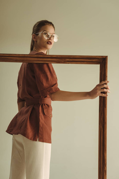 beautiful fashionable girl posing with big wooden frame isolated on beige - Φωτογραφία, εικόνα