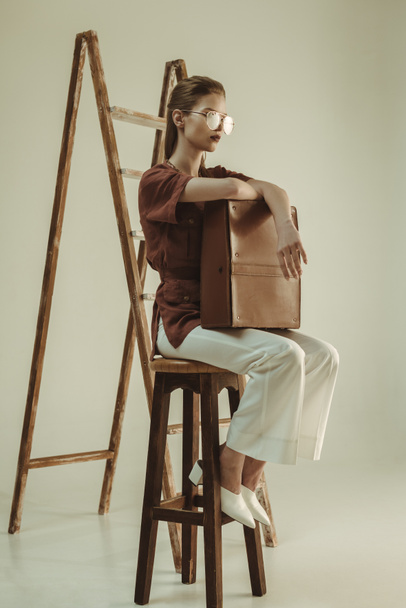 attractive young woman holding leather bag and sitting near ladder on beige - Foto, imagen