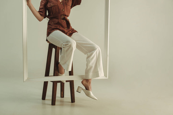 обрезанный вид модной женщины, позирующей на стуле с большой белой рамой
 - Фото, изображение