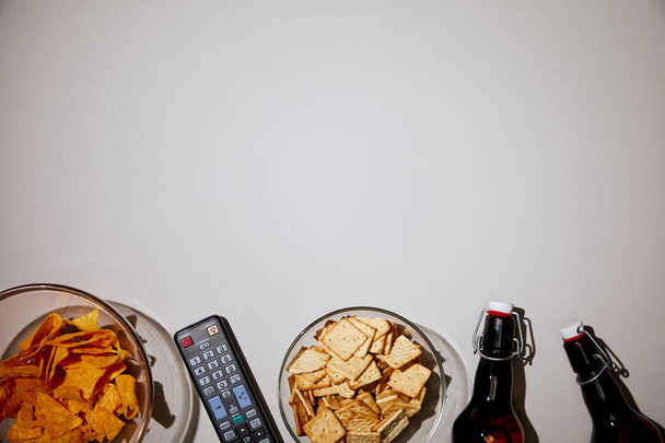 top view of glass bowls with tasty snacks near bottles and remote control on white background - Photo, Image