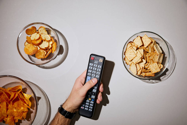 cropped view of man holding remote control in hand near glass bowls with snacks on white background - Φωτογραφία, εικόνα