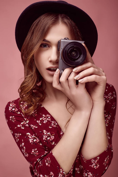 Sensual portrait of elegant glamor hipster girl in red fashion dress, black hat and glasses posing with camera on colorful pink background in studio - Foto, Imagen