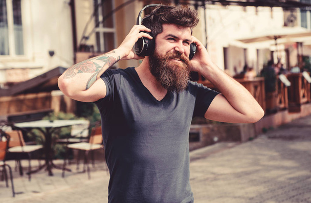 Man with long beard and mustache with wireless headphones on head, defocused urban background. Hipster with headphones on smiling face listening music while walking. Wireless technologies concept - 写真・画像