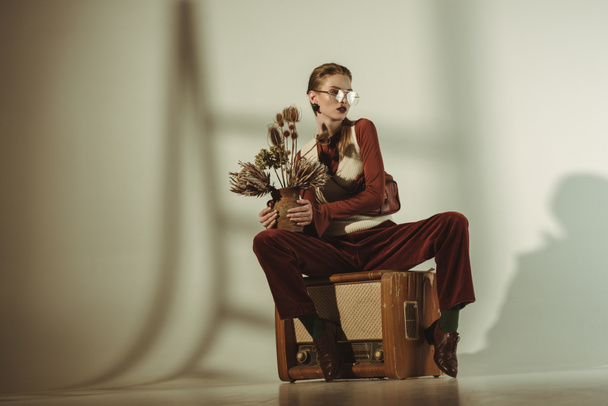 fashionable girl holding bouquet of dry flowers and sitting on vintage tv on beige - 写真・画像