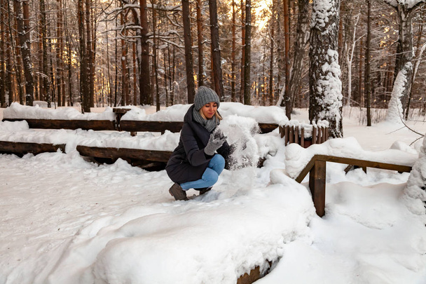 Блондинка в сірому капелюсі і темно-синій піджак взимку в лісі присідає і кидає руки на сніг, граючи з ним, спостерігаючи, як він блищить на сонці
. - Фото, зображення