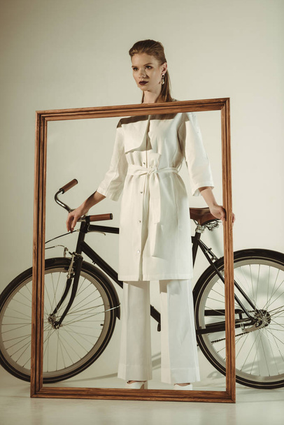 stylish woman in white outfit posing with big wooden frame near bicycle - Photo, Image