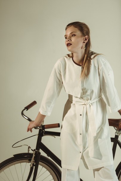 fashionable girl in white outfit posing on bicycle isolated on beige - Фото, изображение