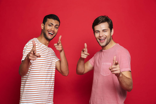 Two cheerful young men standing isolated over red background, pointing at camera - Foto, Bild