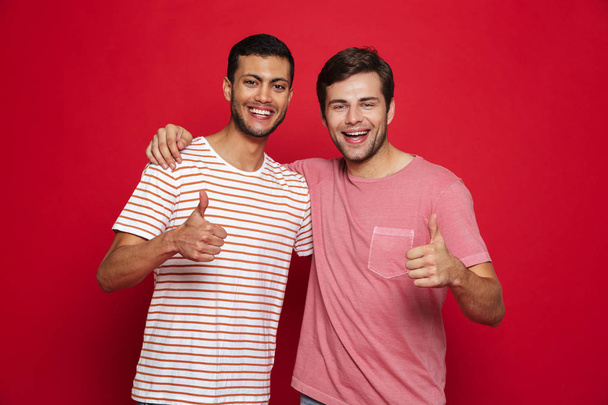 Двоє веселих молодих чоловіків стоять ізольовані на червоному тлі, даючи великі пальці вгору
 - Фото, зображення