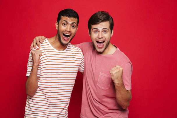 Two cheerful young men standing isolated over red background, celebrating success - 写真・画像