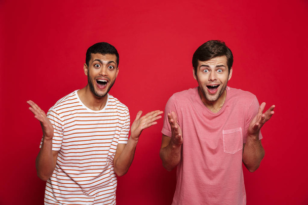 Two cheerful young men standing isolated over red background, celebrating success - Foto, imagen