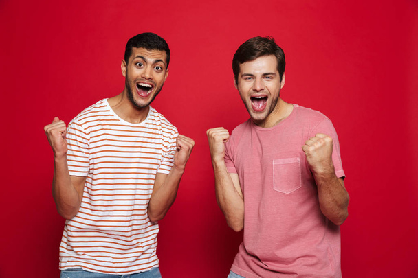 Two cheerful young men standing isolated over red background, celebrating success - Фото, зображення