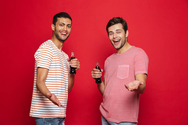 Two cheerful men friends standing isolated over red background, holding bottles with fizzy drink - Φωτογραφία, εικόνα