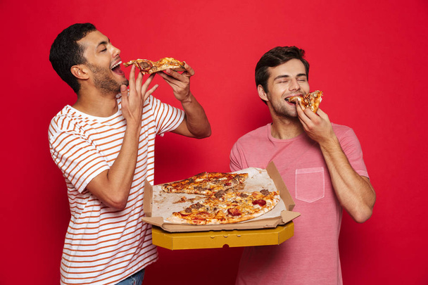 Image of a handsome pleased happy young men friends isolated over red wall background holding big pizza eating. - Photo, Image
