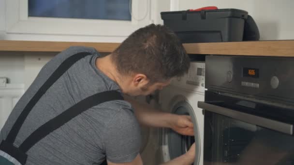 Professional handyman in overalls repairing washing machine in the kitchen. - Footage, Video
