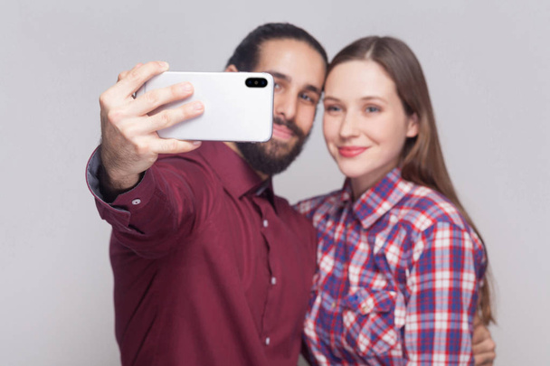 Portrait of happy satisfied couple looking and smiling at smartphone camera to make selfie on grey background.   - Foto, imagen