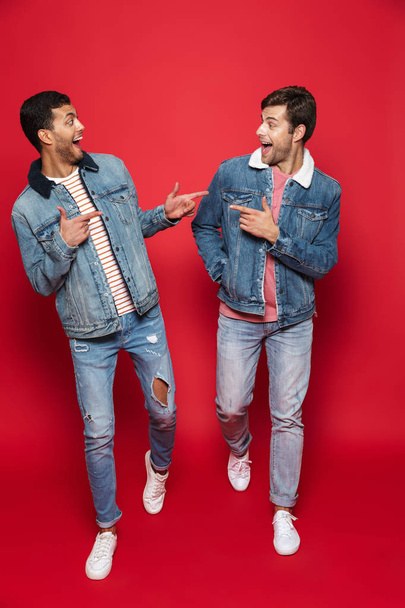 Full length of two excited young men friends wearing denim jackets isolated over red background, walking, talking - Foto, Imagem