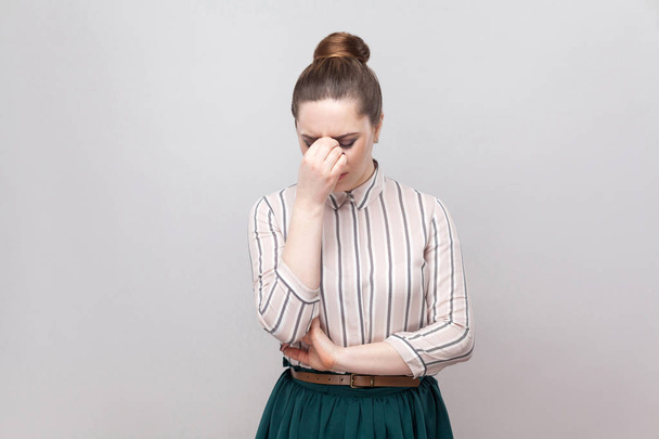 Portrait of sad depressed young woman in striped shirt and green skirt with collected ban hairstyle holding head down and crying  - Photo, image