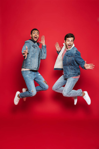 Full length of two excited young men friends wearing denim jackets isolated over red background, jumping - Foto, Imagen