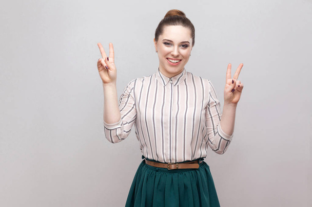 Happy beautiful young woman in striped shirt and green skirt with collected ban hairstyle standing with victory sign   - Photo, image