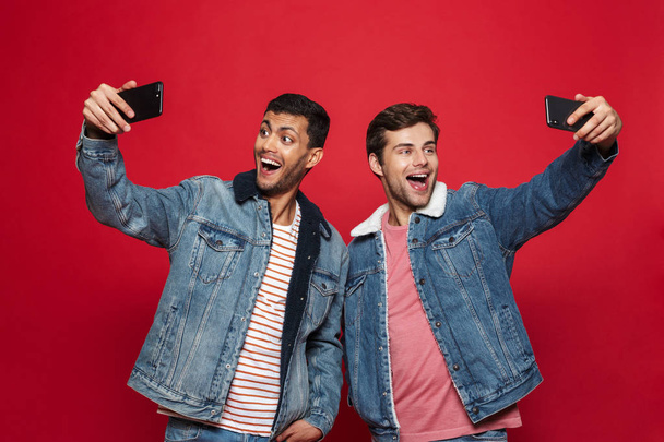 Two cheerful young men standing isolated over red background, taking a selfie - Фото, изображение