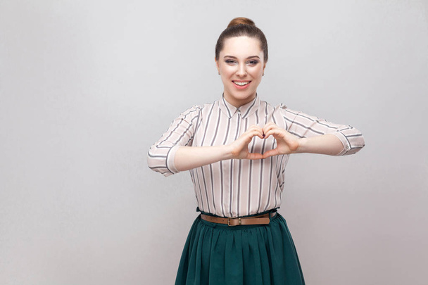 Beautiful young woman in striped shirt and green skirt with ban hairstyle standing with heart gesture and looking at camera  - Fotografie, Obrázek