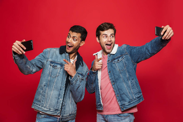 Two cheerful young men standing isolated over red background, taking a selfie - Φωτογραφία, εικόνα