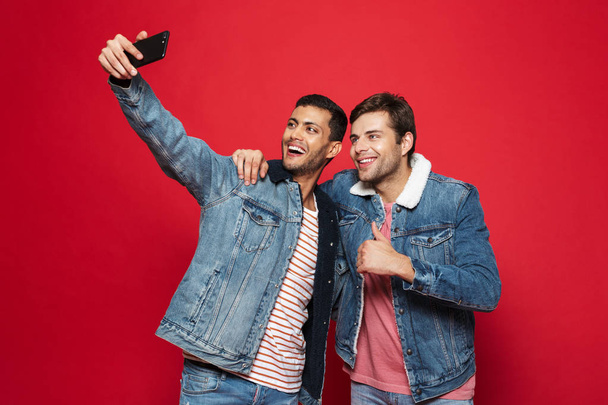 Two cheerful young men standing isolated over red background, taking a selfie - Photo, Image