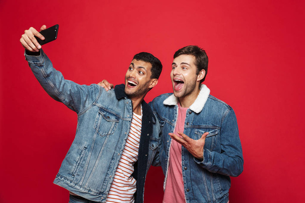 Two cheerful young men standing isolated over red background, taking a selfie - Zdjęcie, obraz