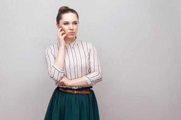 Confused woman in striped shirt and green skirt with makeup and collected ban hairstyle standing with thoughtful face and looking away - 写真・画像
