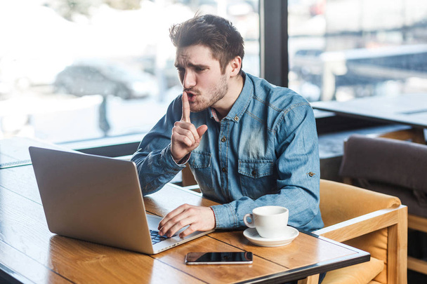 Portrait of handsome severe bearded freelancer in blue jeans shirt leaning finger on mouth and showing silence sign while sitting with laptop in cafe   - Foto, Imagen