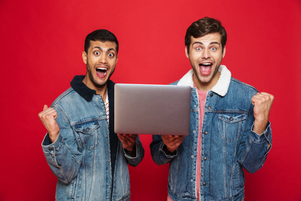 Two cheerful men friends standing isolated over red background, holding laptop computer, celebrating - Фото, зображення