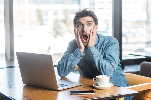 Portrait of emotional scared young businessman in blue jeans shirt with raised arms and stressed face looking at camera while sitting with laptop and screaming in cafe  - Photo, image