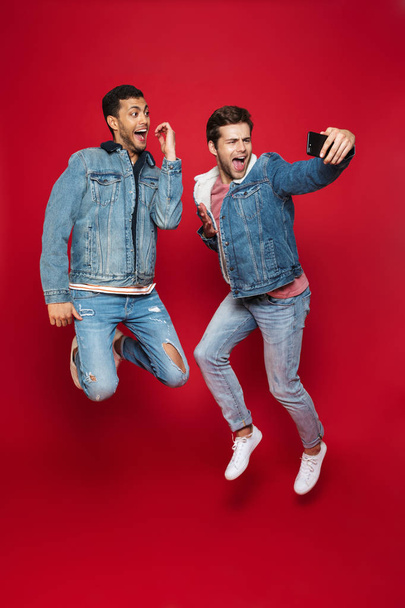Two cheerful young men jumping isolated over red background, taking a selfie - Foto, Imagen