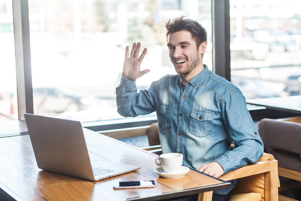 Portrait of satisfied happy young businessman in blue jeans shirt sitting in cafe and greeting worker through webcam while showing palm of hand gesture with toothy smile. - Фото, изображение
