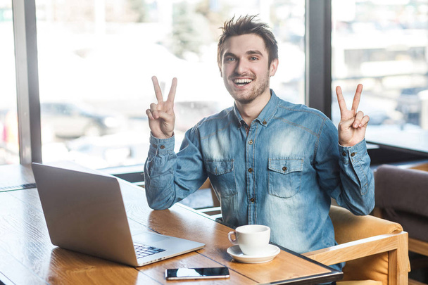 Portrait of positive bearded freelancer in blue jeans shirt sitting in cafe and working on laptop with toothy smile while showing victory sign at camera  - Photo, Image