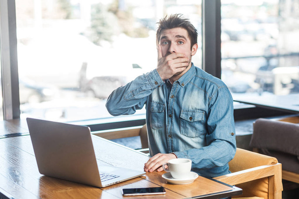 portrait of emotional scared businessman in blue jeans shirt sitting in cafe with laptop and covering mouth with hand while looking at camera  - Fotoğraf, Görsel