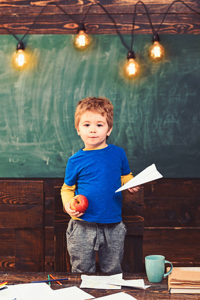 Little boy holding paper plane and apple in his hands. Schoolboy in blue T-shirt standing behind the table. Educational game concept - Foto, Imagem