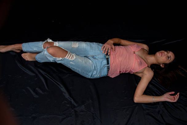 Girl with pink tank top in transparent lace on chest and torn fashionable blue jeans. Full length of young girl with long hair, lying on black background. Sexy female lingerie. Copy space. - Foto, Imagem