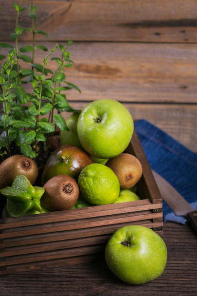 Healthy food background. Concept of healthy food. Fresh green fruits in box on dark wooden background. Set of green fruits for healthy diet and detox: apple, lime, kiwi, mango, carambola and mint. - Фото, изображение