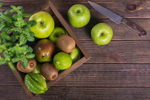Concept of healthy food. Fresh green fruits in box on dark wooden background. Set of green fruits for healthy diet and detox: apple, lime, kiwi, mango, carambola and mint. Flat lay - Photo, Image