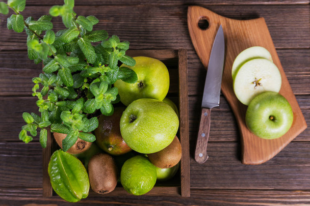 Concept of healthy food. Fresh green fruits in a box, green apple on cutting board. Set of green fruits for healthy diet and detox: apple, lime, kiwi, mango, carambola and mint. top view - Foto, Imagem