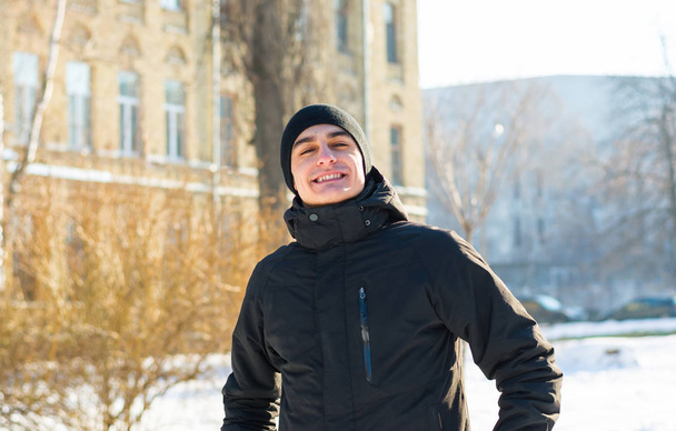 Egy fiatal srác téli ruhákat, házak, télen a háttérben. Egy diák áll, és az egyetemi épületek télen mosolyog - Fotó, kép