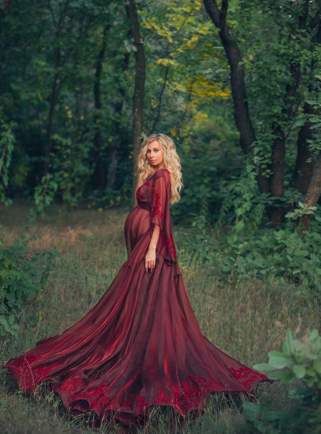 young pregnant woman with blond curly hair in long light flapping red scarlet dress, standing in the forest, holding, hugging her tummy. like a fairy magnificent queen. art autumn dark halloween photo - Foto, imagen