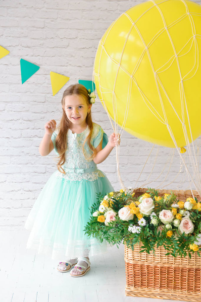 A girl in an elegant dress is standing in the studio next to a large balloon and a basket of flowers. Festive decor, birthday decorations - 写真・画像