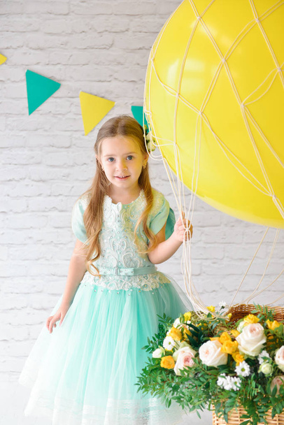 A girl in an elegant dress is standing in the studio next to a large balloon and a basket of flowers. Festive decor, birthday decorations - Photo, Image