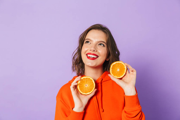 Image of an excited beautiful young woman posing isolated over purple background wall holding orange citrus. - Zdjęcie, obraz