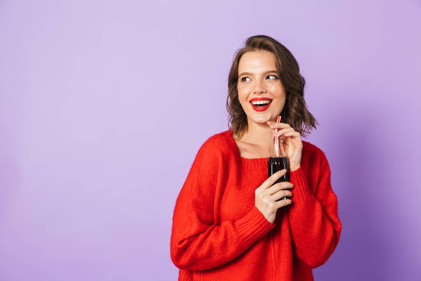 Excited young girl wearing red sweater standing isolated over violet background, drinking from a glass bottle with a straw - Fotó, kép